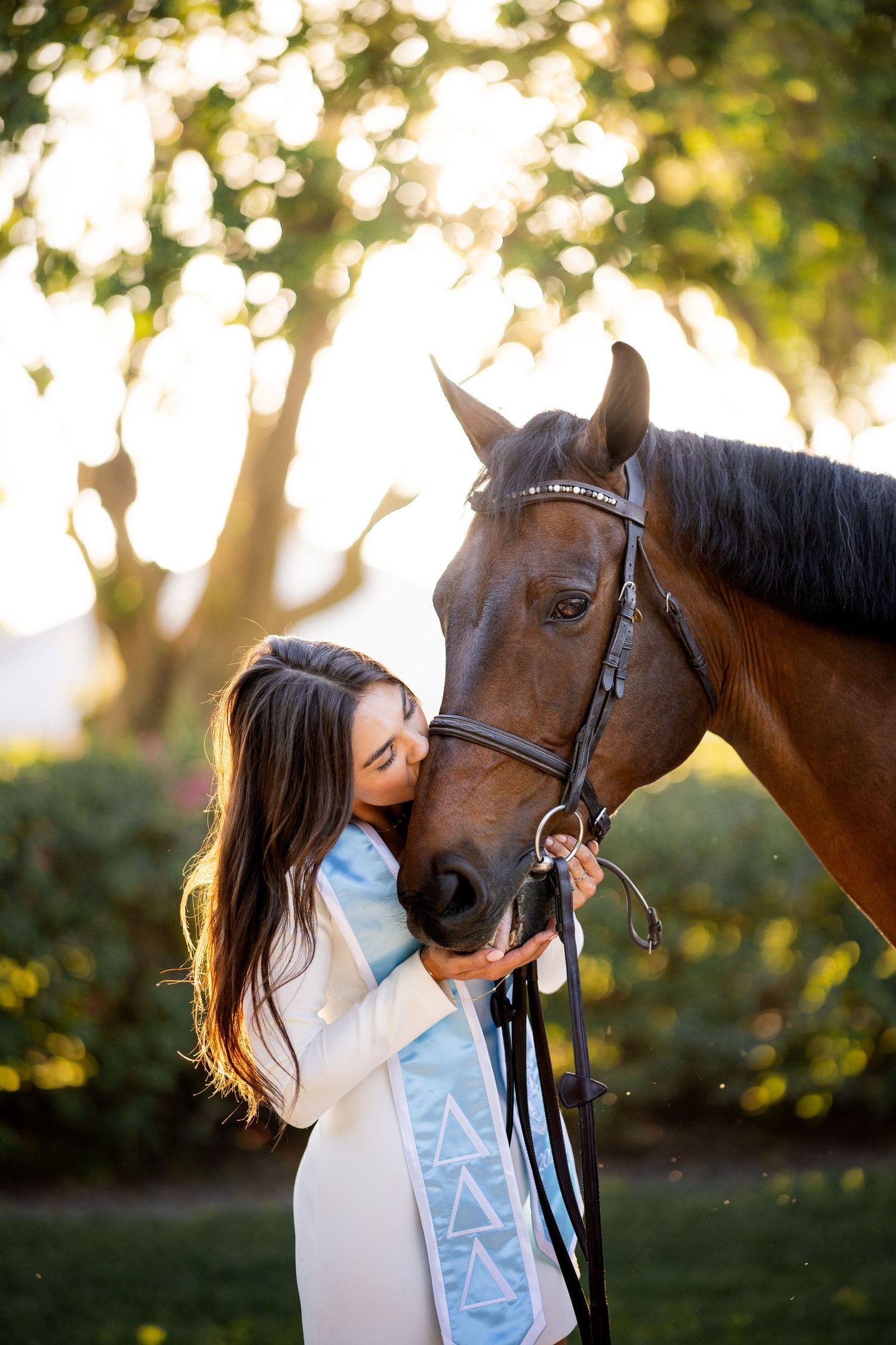 Young woman in graduation sash kissing a brown horse outdoors in golden sunlight with lush green trees.