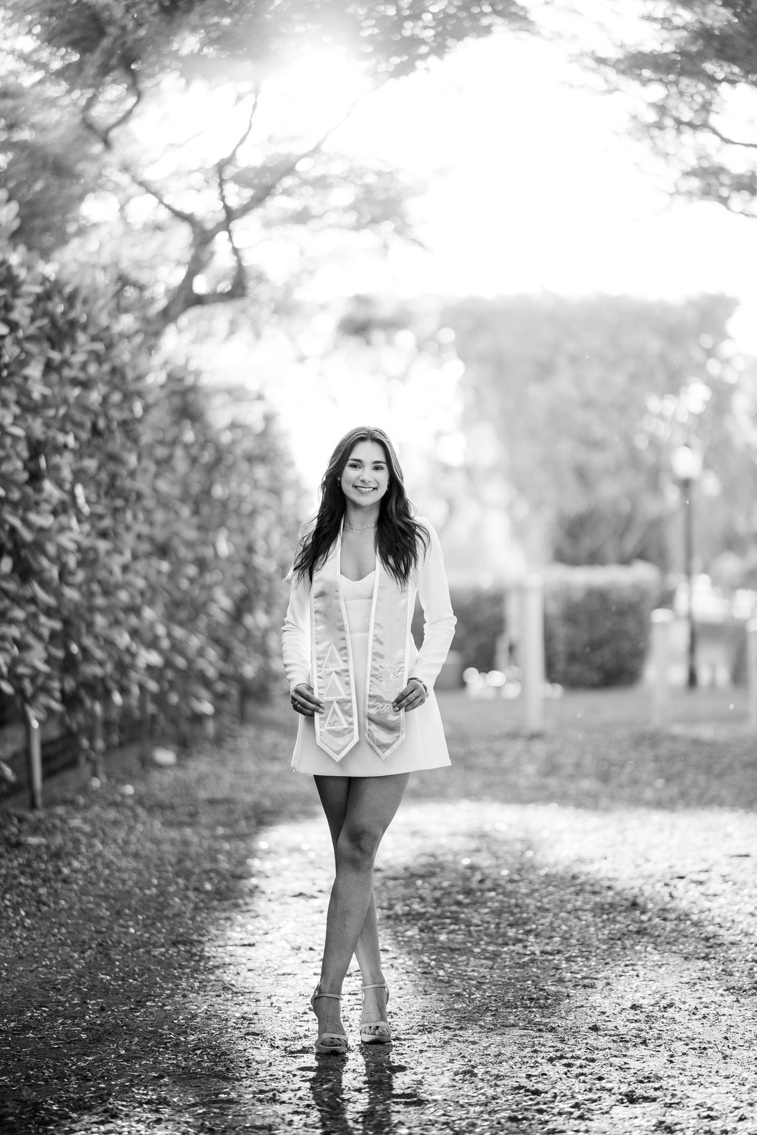 Black and white photo of a smiling young woman in a short white dress walking down a tree-lined path.