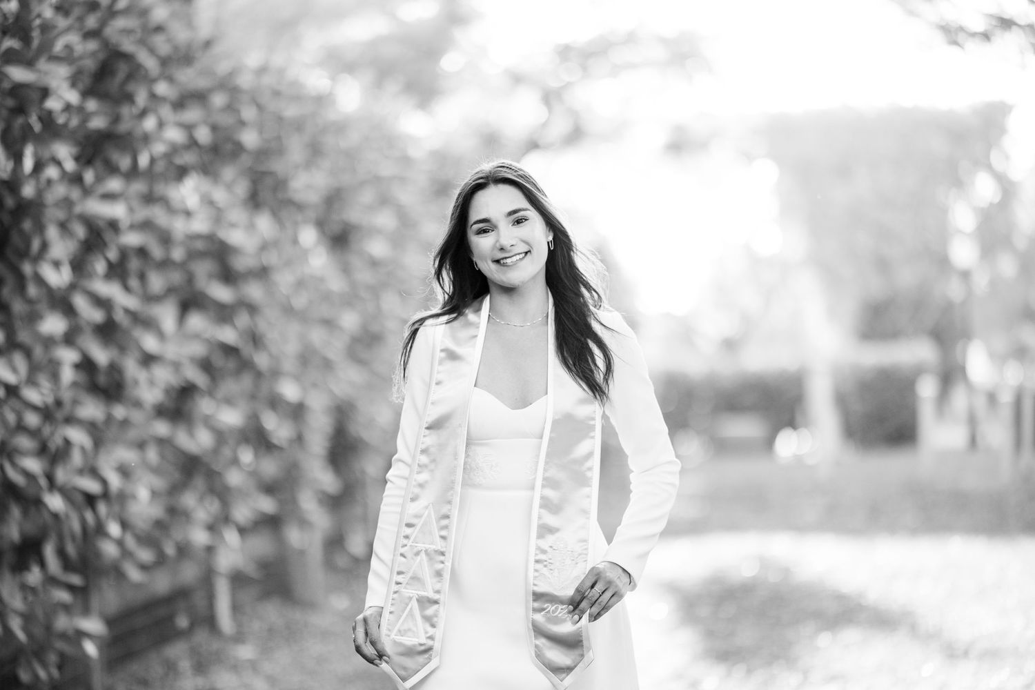 Black and white photo of a smiling woman in a white dress and jacket posing outdoors in a lush garden setting.