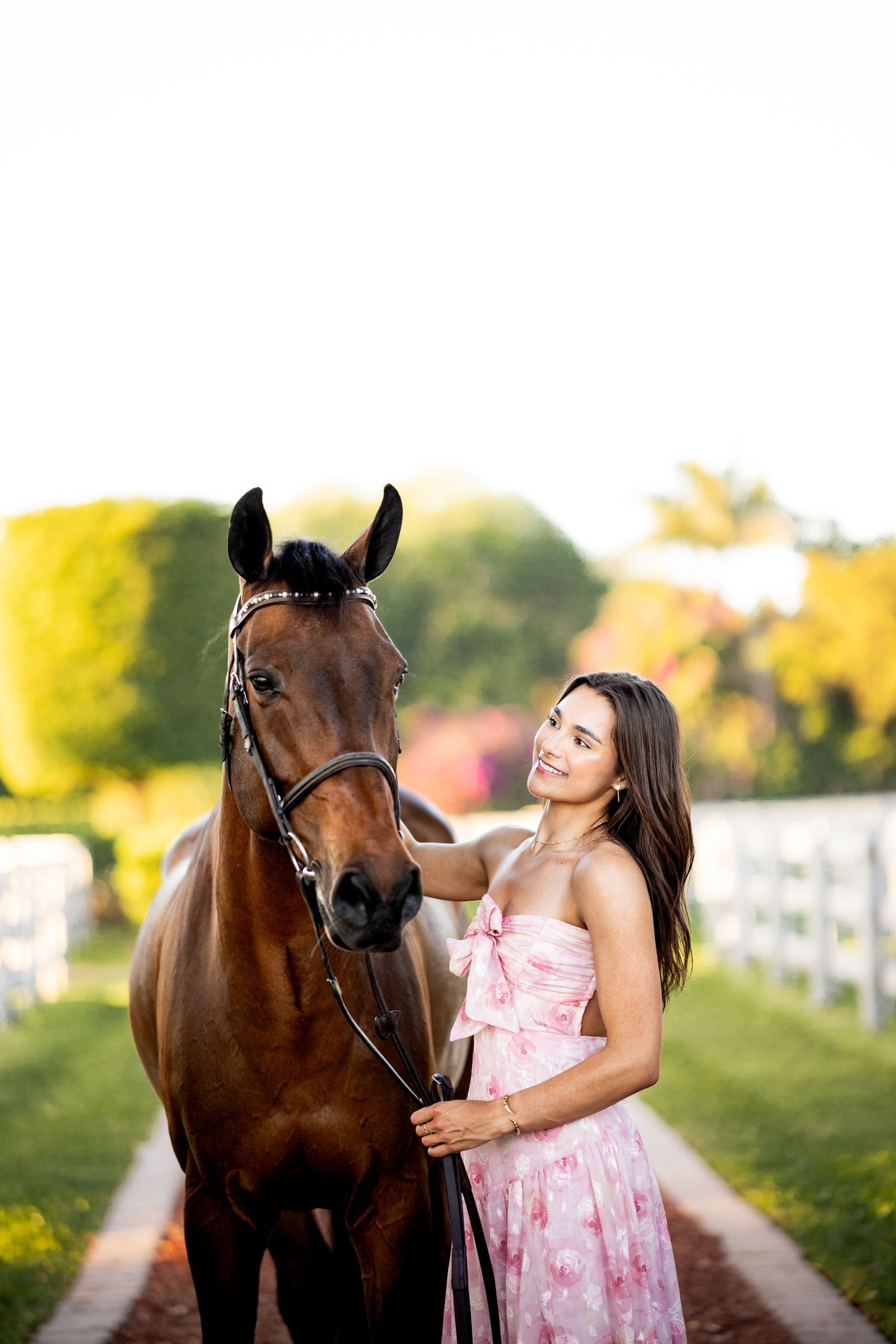 A young woman in a pink floral dress smiles while standing beside a bay horse on a sunny green pathway.