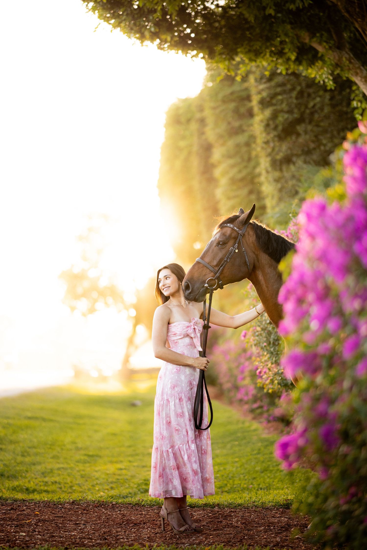 Young woman in pink floral dress stands with brown horse amid purple flowers and golden sunset light.