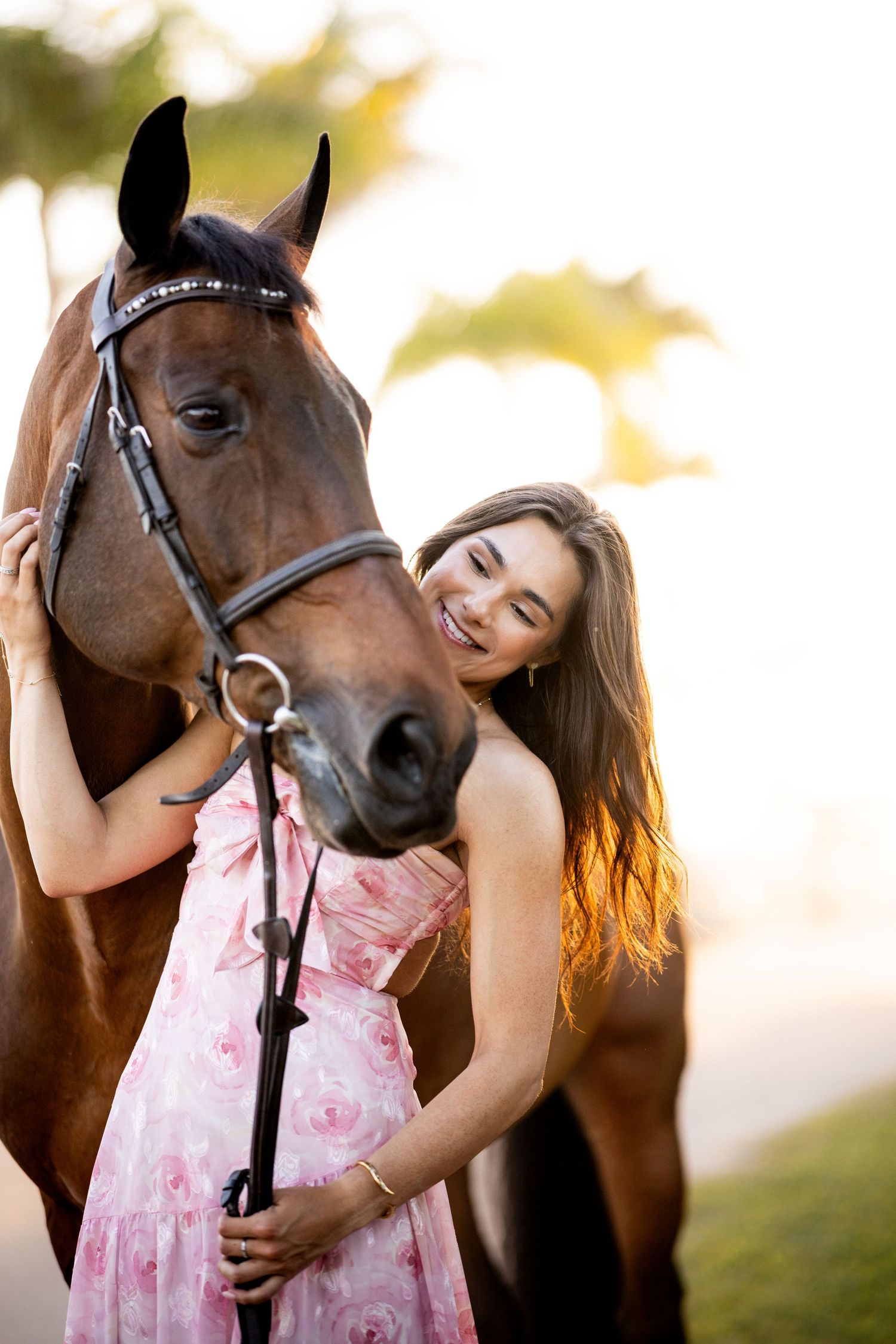 Smiling young woman in pink floral dress affectionately posing with a bay horse outdoors at golden hour.