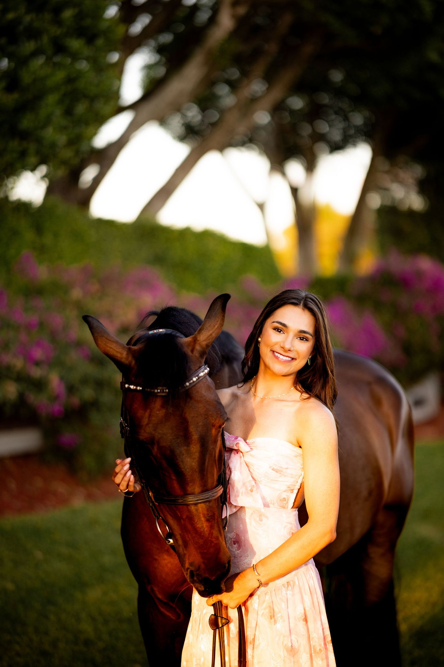 Young woman in floral dress smiling beside a dark brown horse in a lush garden with purple flowers.
