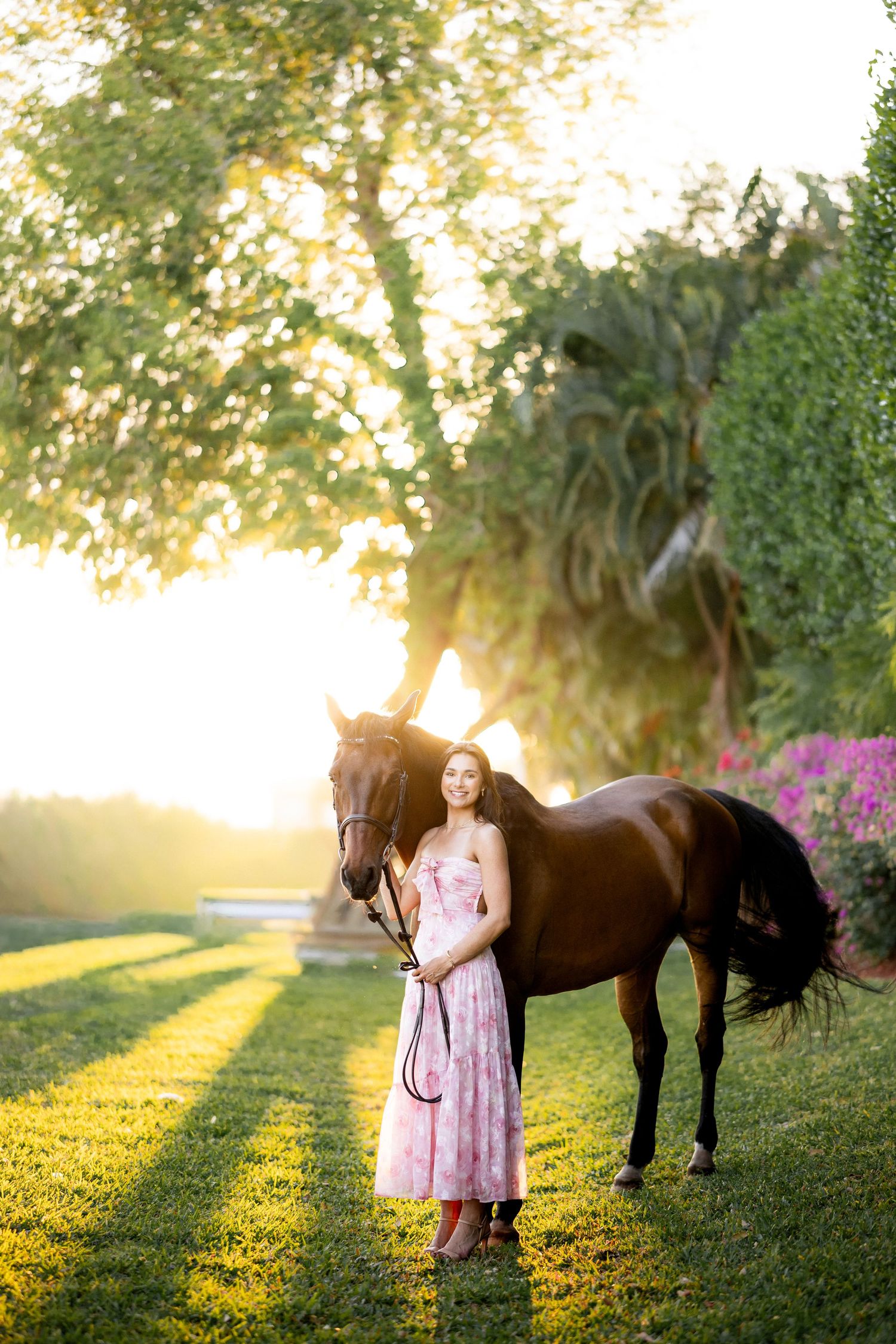 Young woman in pink floral dress stands with dark bay horse at golden hour in lush green garden setting.