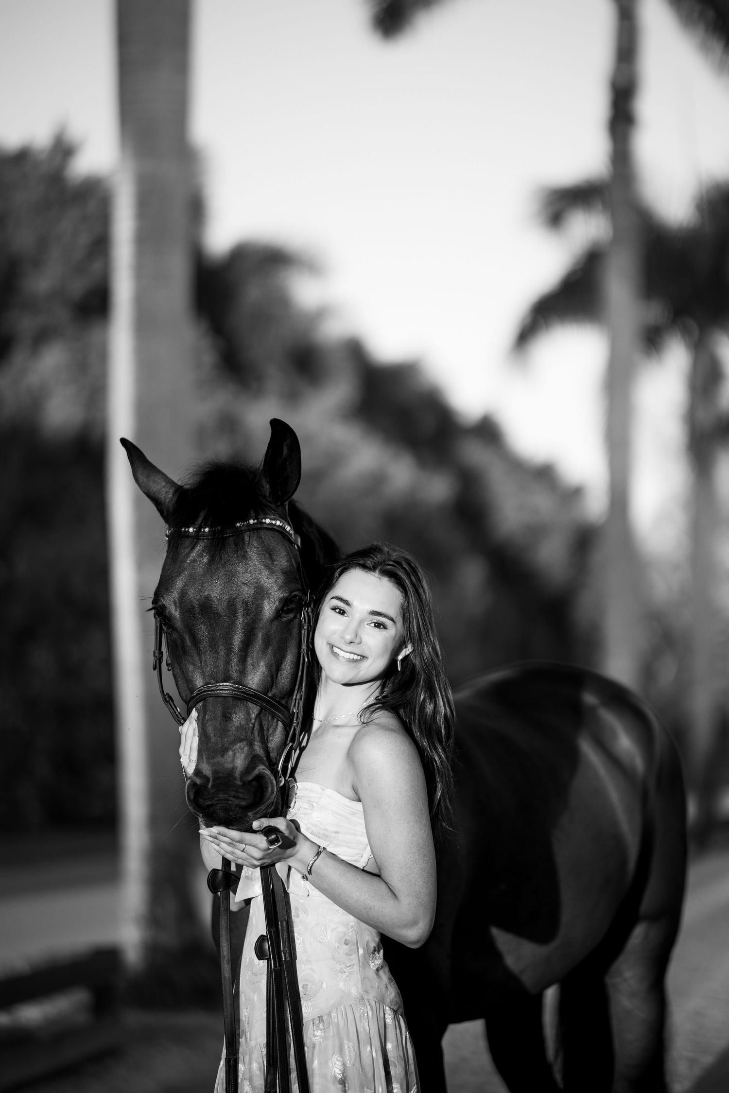 Black and white photo of a smiling young woman in a dress posing with a dark horse, palm trees in background.