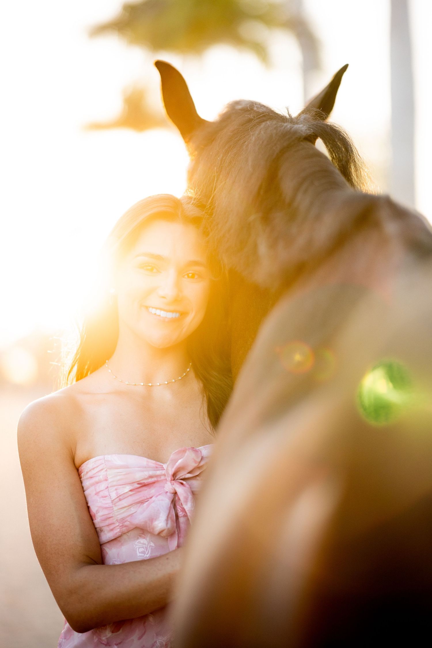 A smiling young woman in a pink strapless dress stands beside a horse in warm golden sunset light.