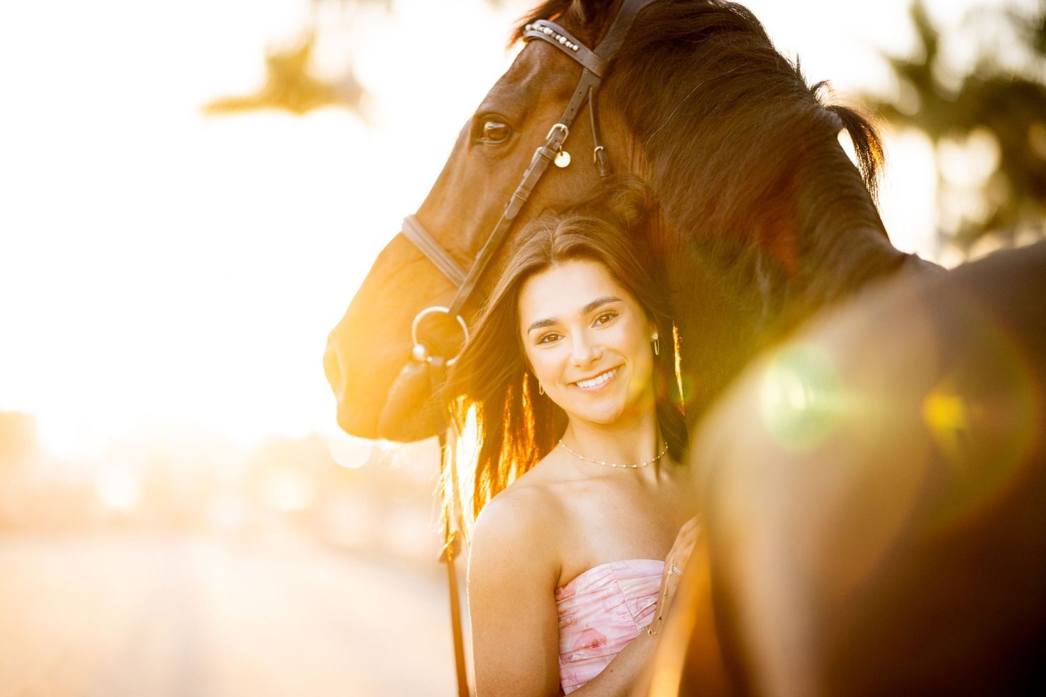 Young woman in pink dress smiling beside a dark horse at golden hour sunset.