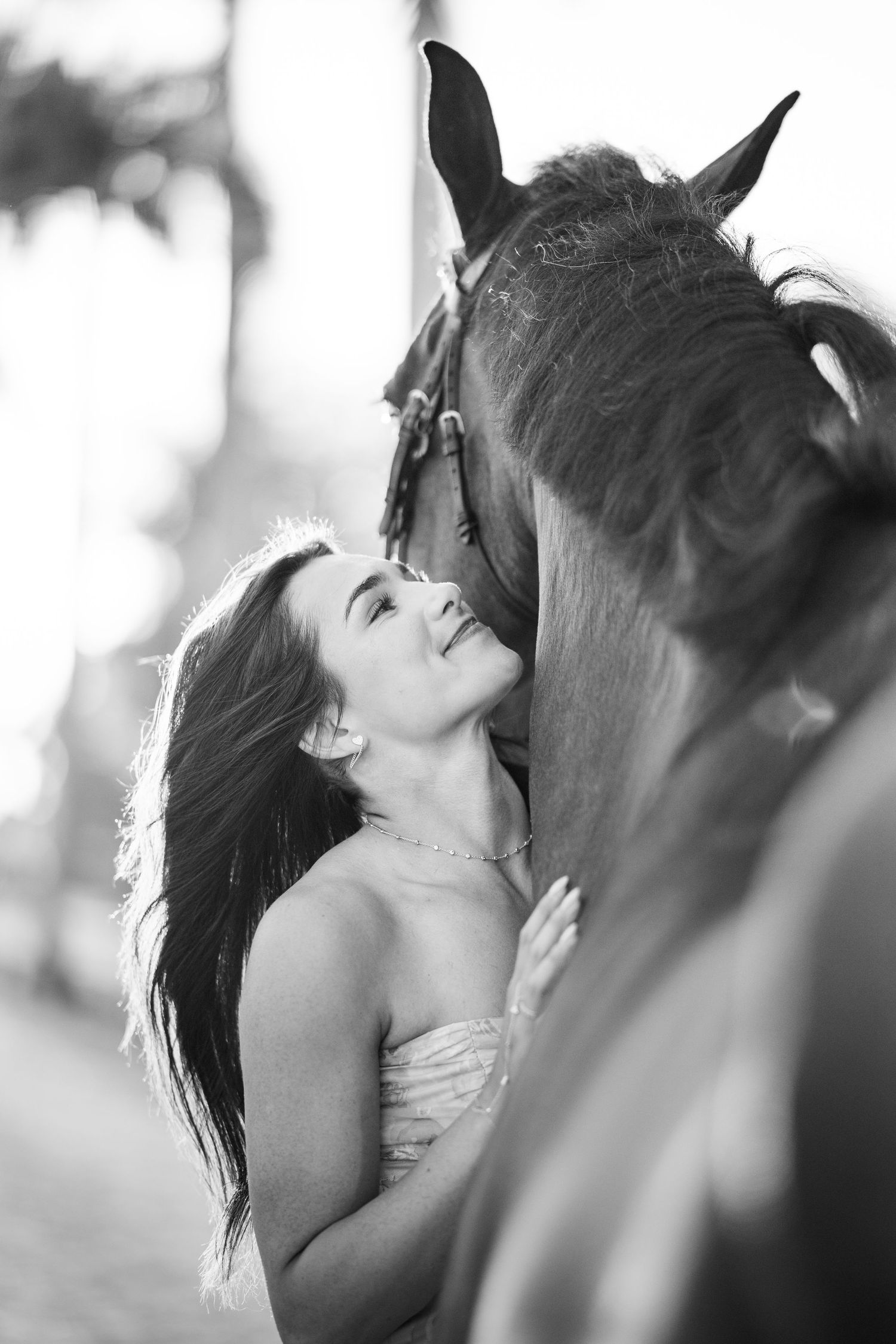 Black and white photo of a woman gazing up tenderly at a dark horse outdoors.
