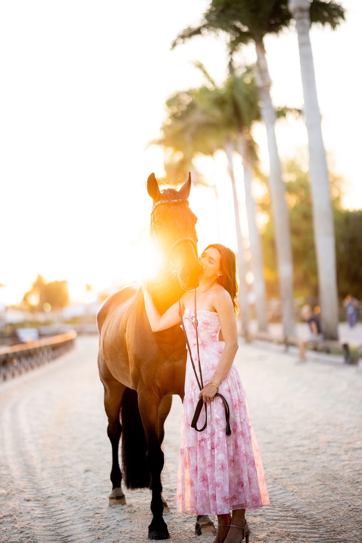 Woman in pink floral dress stands with bay horse on cobblestone path framed by palm trees at golden hour.