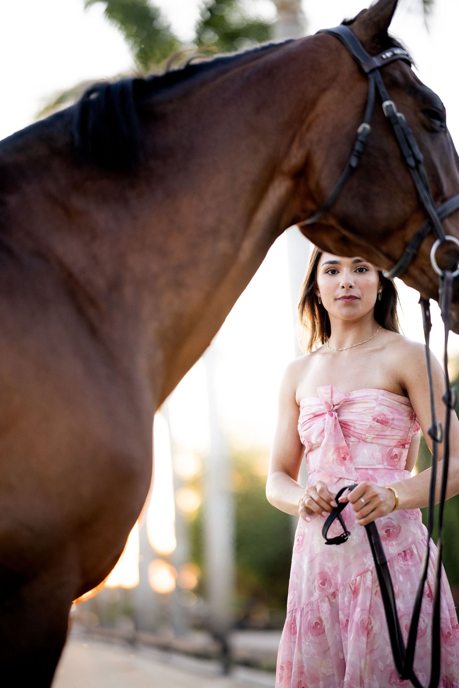Young woman in pink floral strapless dress holding reins beside a brown horse outdoors.