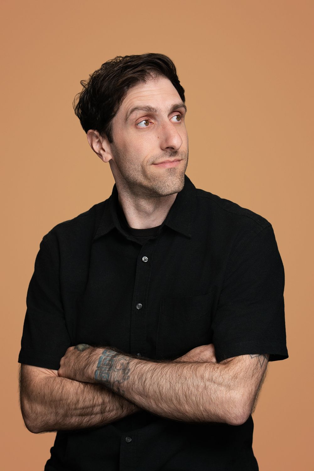 Man in black shirt with crossed arms and tattoo, smirking against a warm beige background.