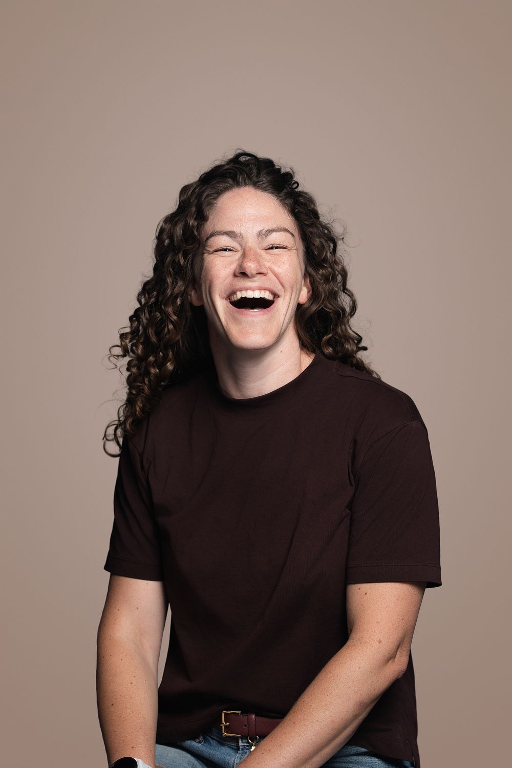 A young woman with curly hair laughs joyfully wearing a dark brown t-shirt against a warm beige background.