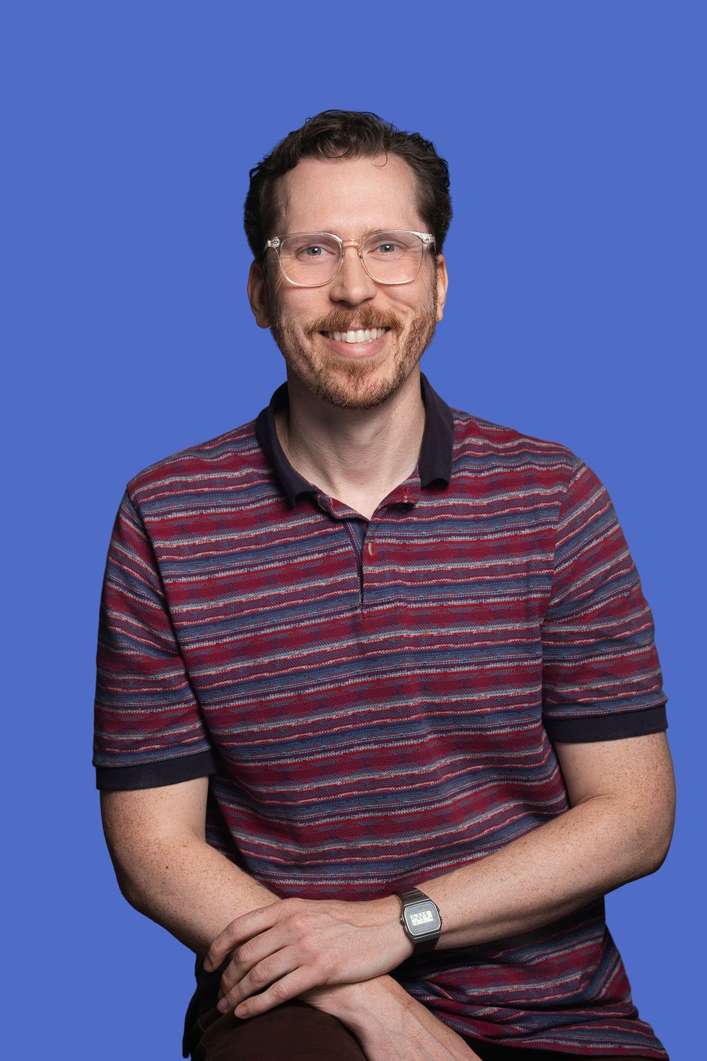 Smiling man with glasses and beard wearing striped polo shirt, seated against blue background.