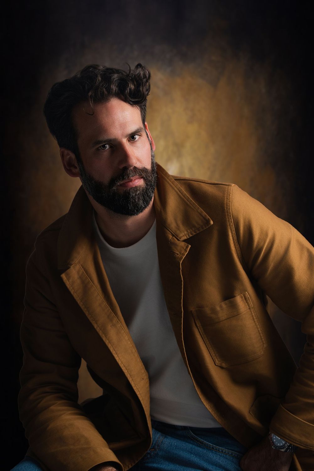 Bearded man in a tan jacket and gray shirt poses against a dark golden studio background.