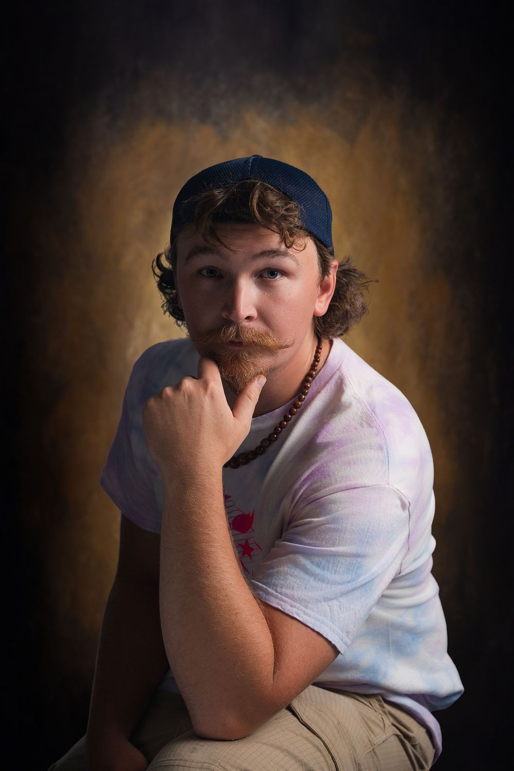 Young man with mustache wearing backward blue cap and tie-dye shirt poses thoughtfully against dark studio backdrop.