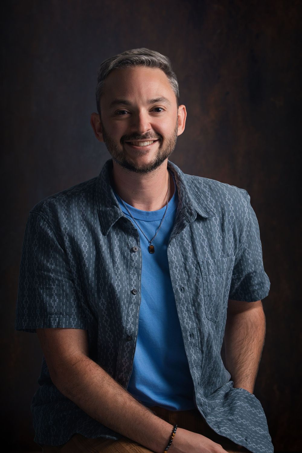 Smiling man with gray hair and beard wearing a blue patterned shirt over a blue tee, seated against a dark background.