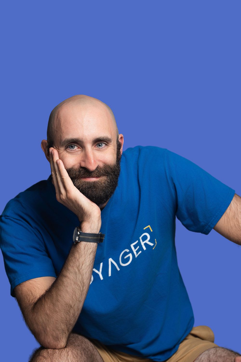 Bald bearded man in blue Voyager t-shirt poses with hand on cheek against purple background.