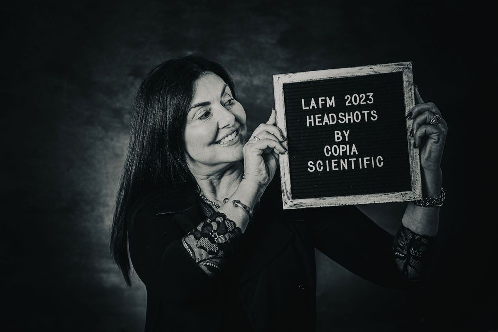 Black and white headshot photo of a smiling woman holding a letterboard sign reading LAFM 2023 Headshots by Copia Scientific.