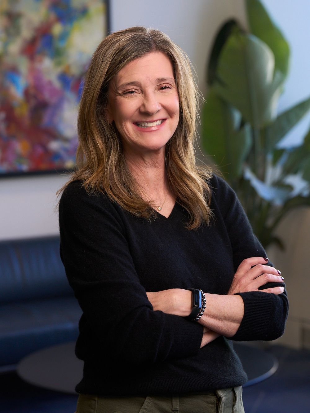 A smiling woman with long brown hair wearing a black sweater stands with arms crossed in a modern office.