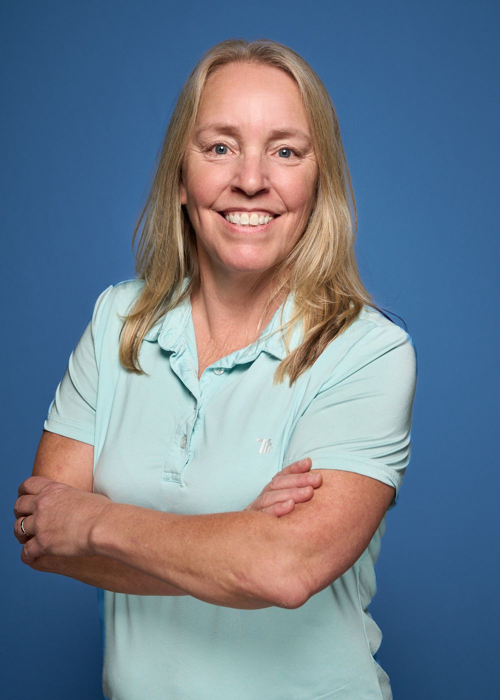 Woman with blonde hair in light blue polo shirt smiling with arms crossed against blue background.