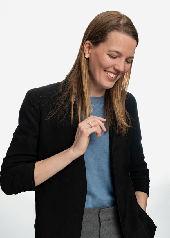 Smiling woman in black blazer and blue top with long brown hair, wearing gold jewelry, on white background.