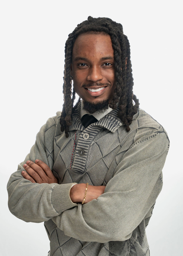 Smiling young man with dreadlocks, crossed arms, wearing a gray cable-knit sweater, posing for a professional portrait.