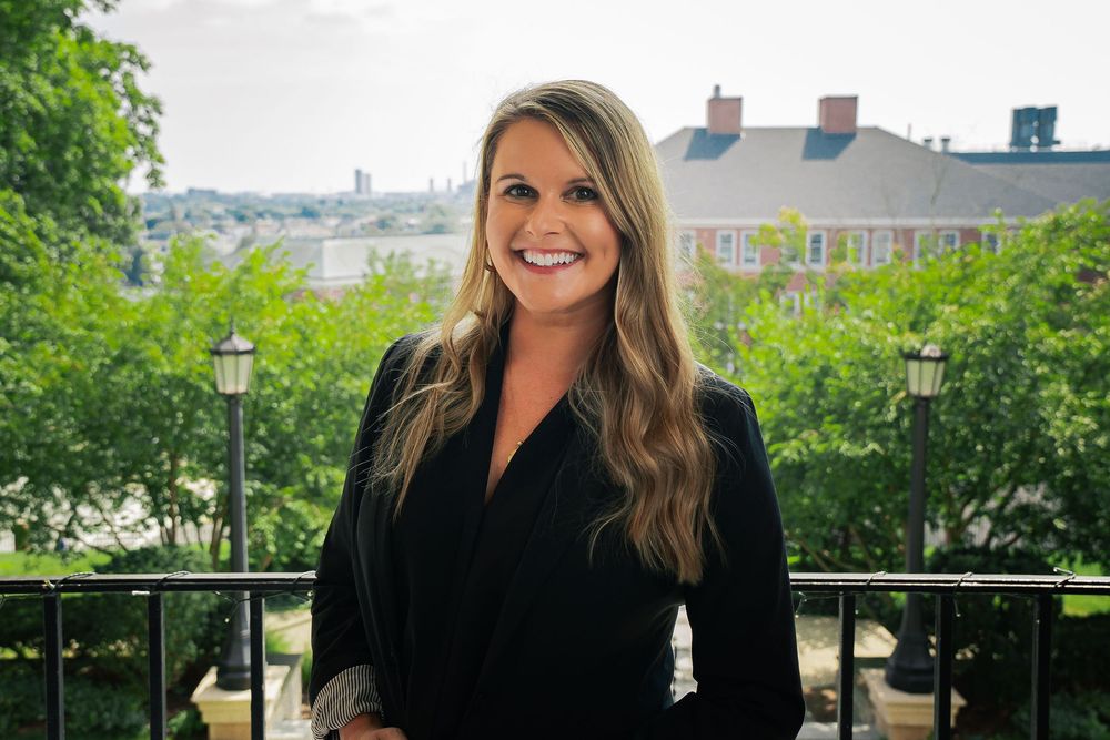 Smiling blonde woman in black blazer poses on outdoor balcony with lush green trees and historic brick building behind her.