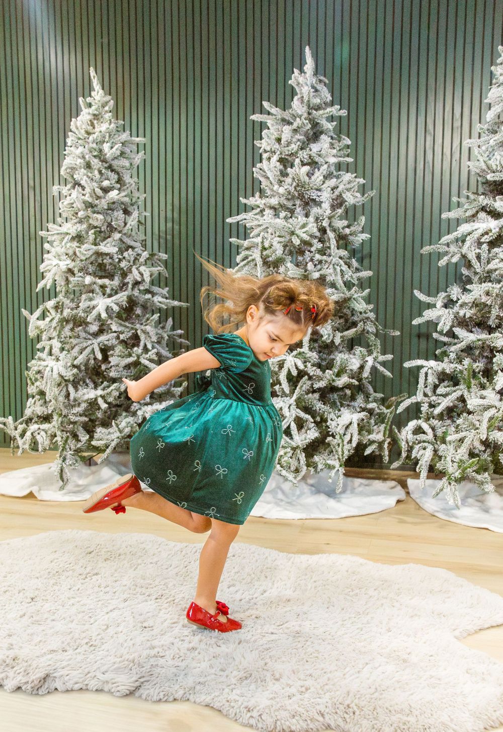 A young girl in a green snowflake dress and red shoes twirls joyfully in front of snow-flocked Christmas trees.