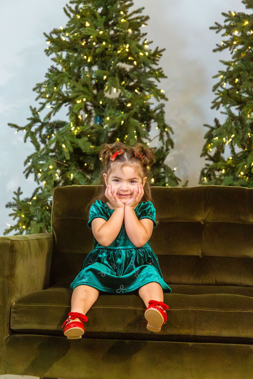 Smiling little girl in green velvet dress sitting on sofa with Christmas trees and lights in background.