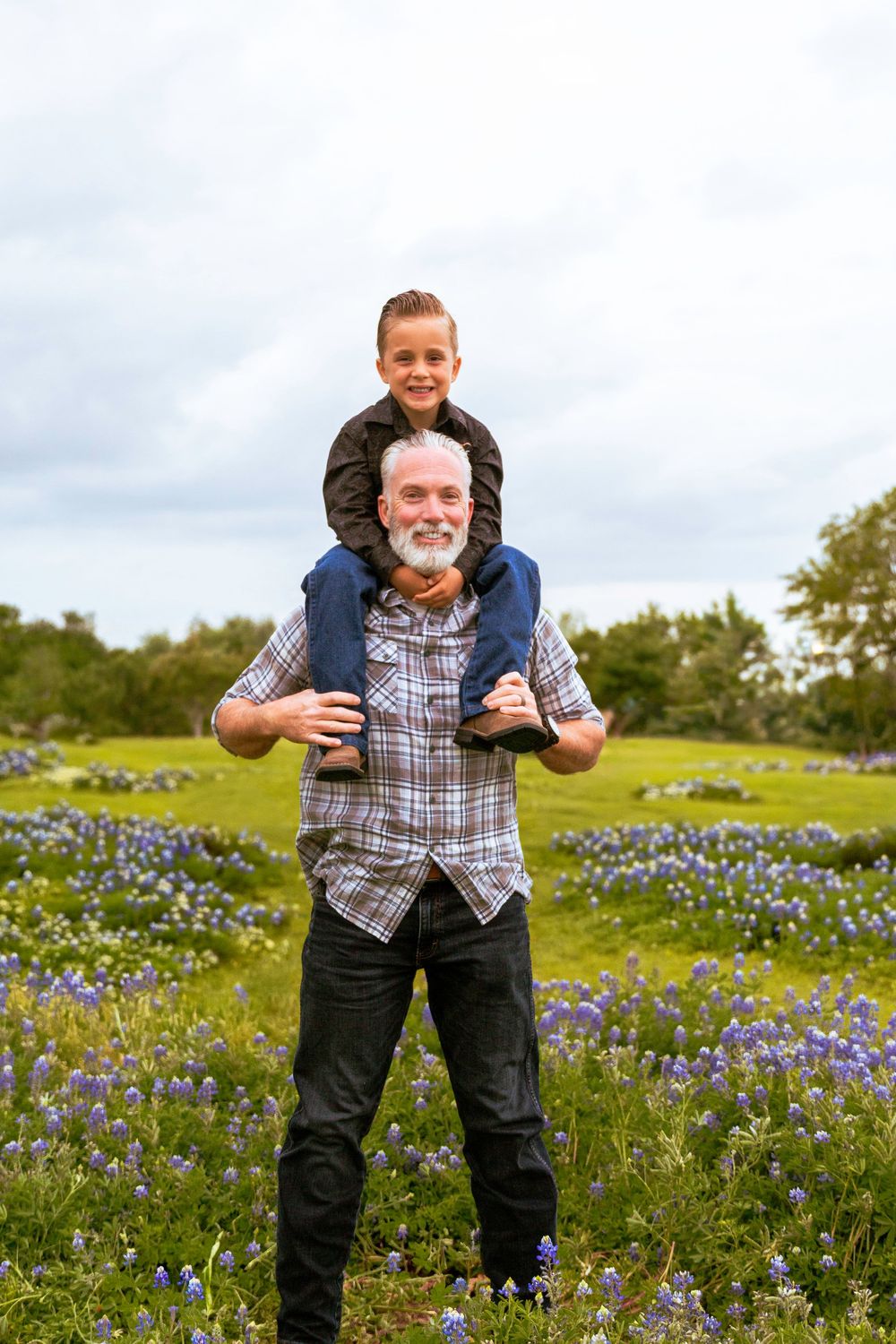 An older man with a gray beard carries a smiling boy on his shoulders in a field of blooming bluebonnets.