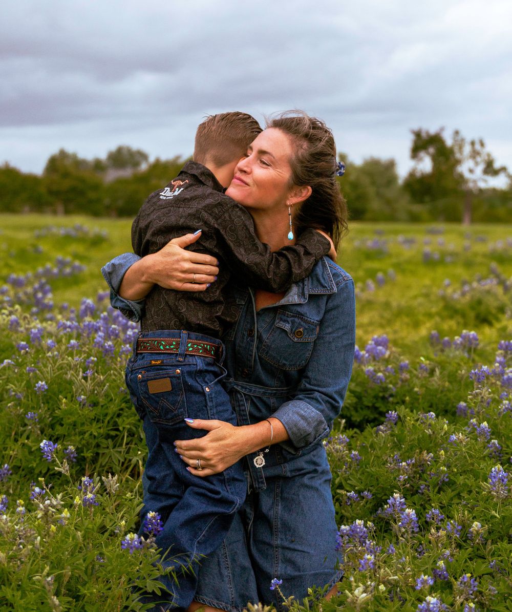 A woman hugs a young boy in a field of purple wildflowers under a cloudy sky.