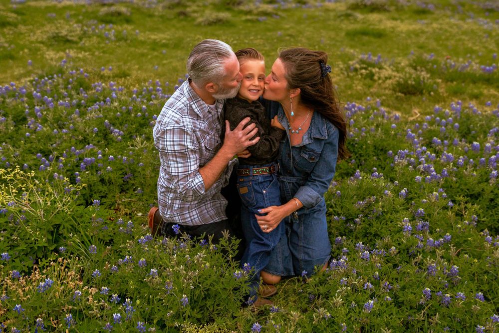 Family of three sharing kisses in a blooming wildflower field, parents embracing child surrounded by purple lupines.