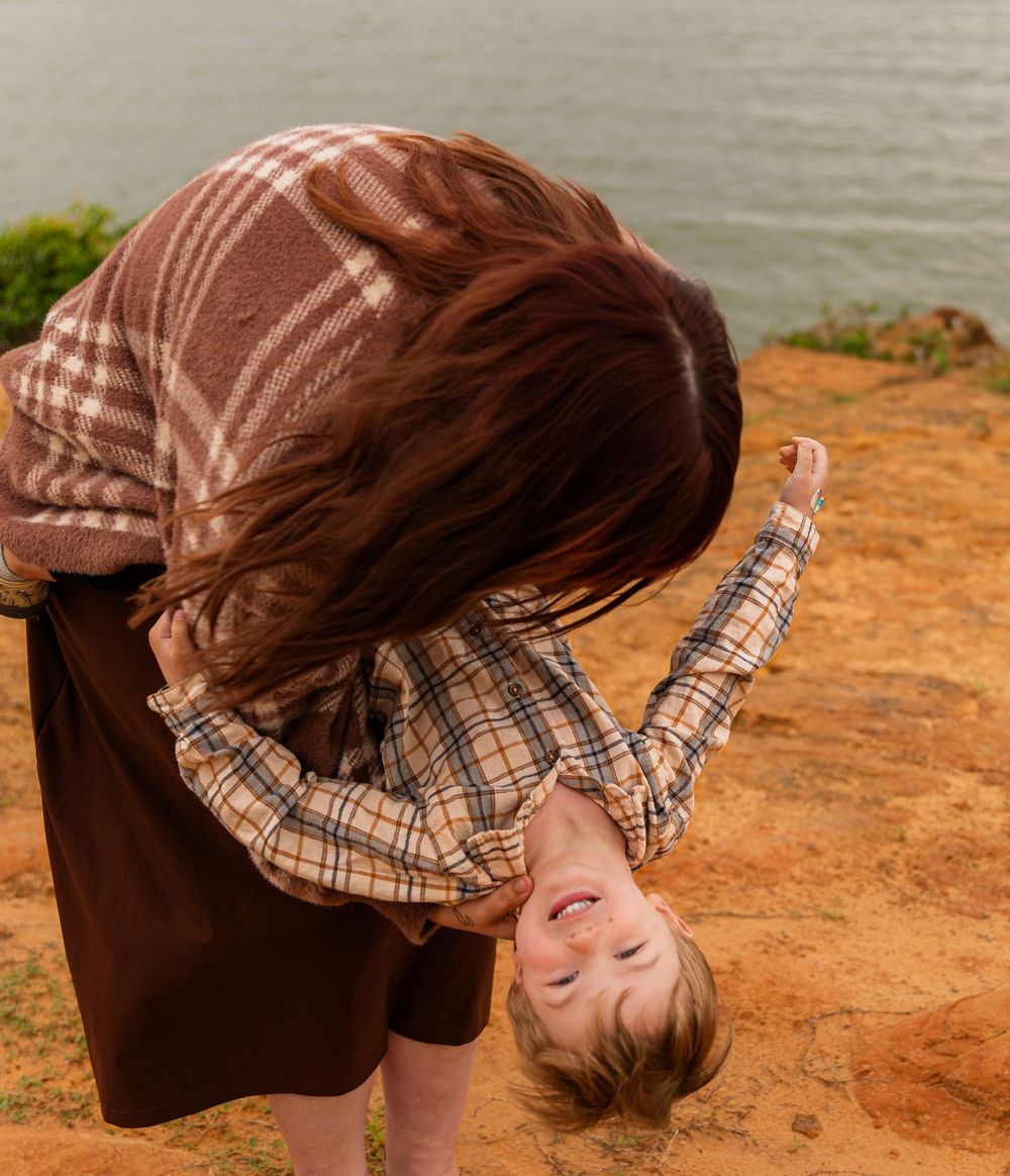 A mother playfully holds her laughing child upside down near a scenic waterfront on a rocky cliff.
