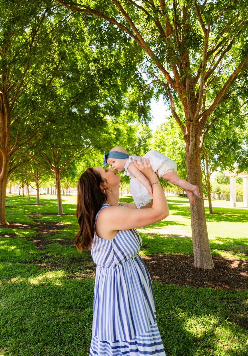 A mother in a blue striped dress lifts her baby up for a kiss in a lush green park.