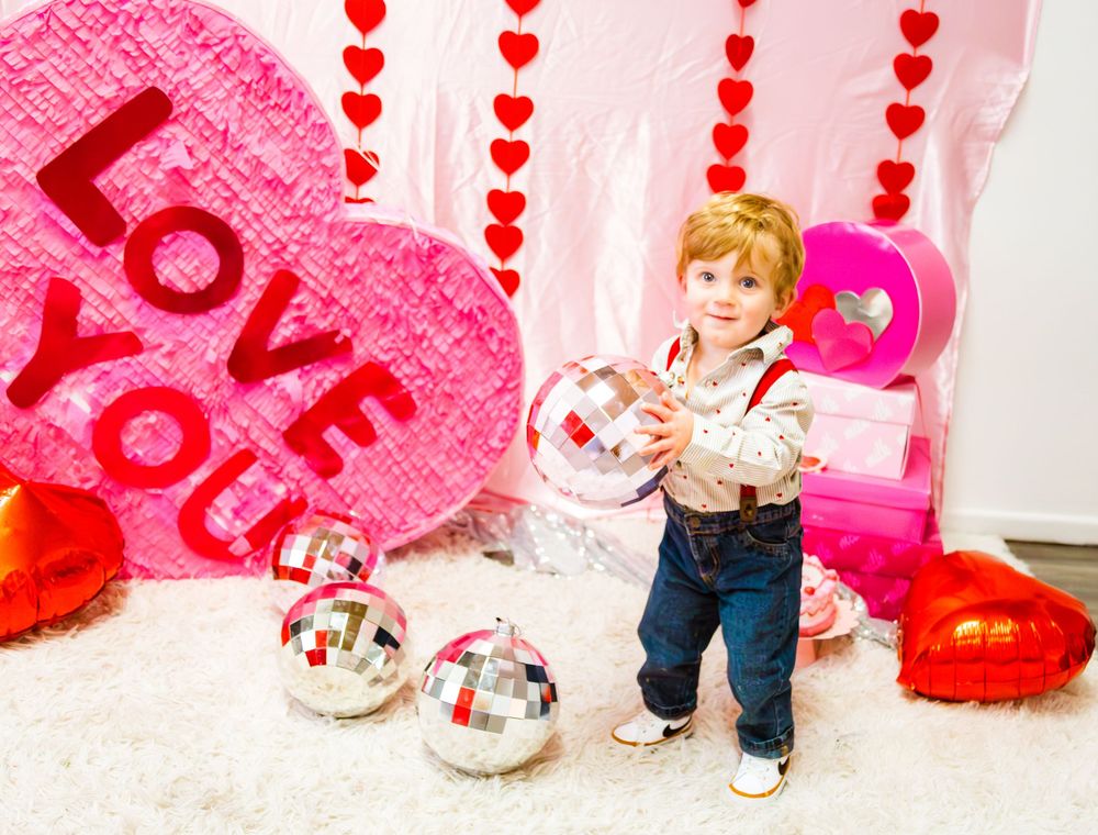 Toddler holding disco ball amid Valentine's Day decorations with Love You heart balloon and red hearts backdrop.