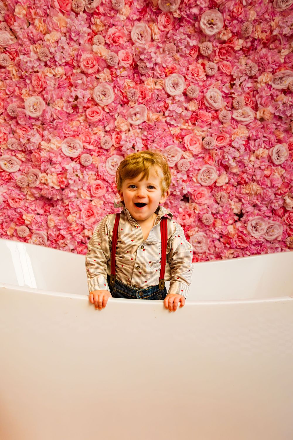 Smiling toddler in suspenders standing in a white bathtub against a pink floral wall backdrop.