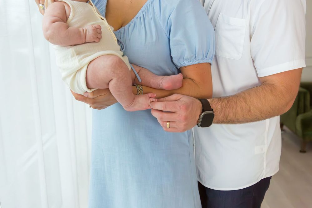 Parents holding a newborn baby together, mother in blue dress and father in white shirt, indoor family portrait.