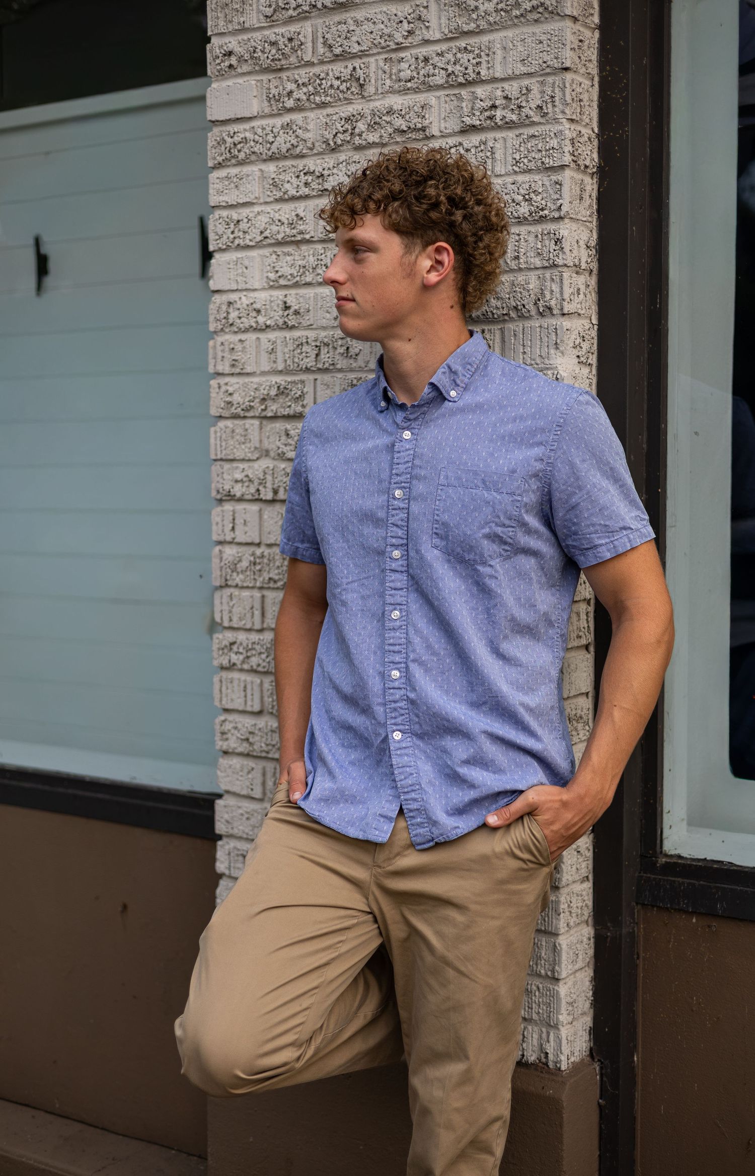 Stylish young man in a blue button-up shirt leaning against a brick wall outdoors.