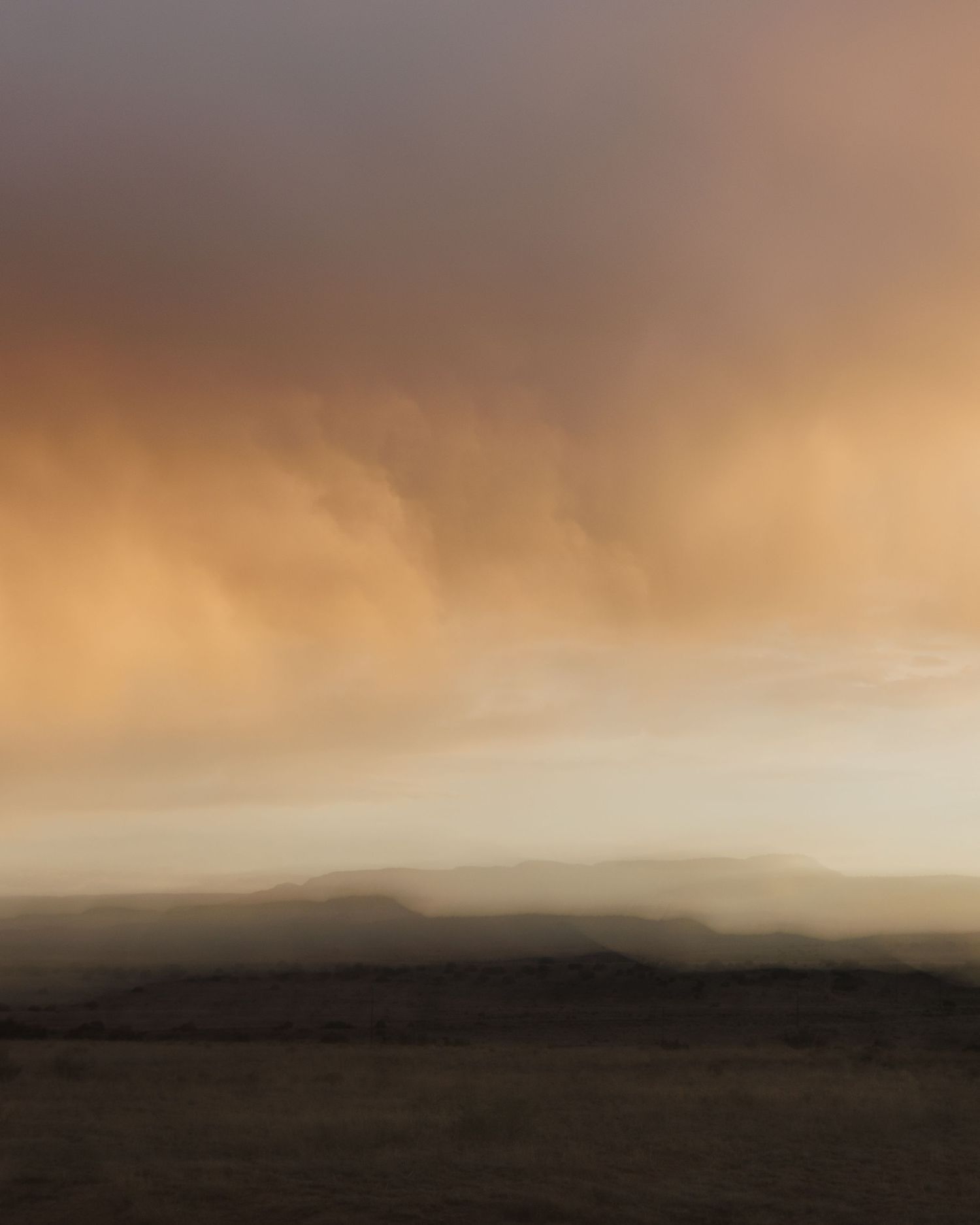 Misty mountain landscape at sunset with soft orange and gray hues enveloping a silhouetted peak in fog.