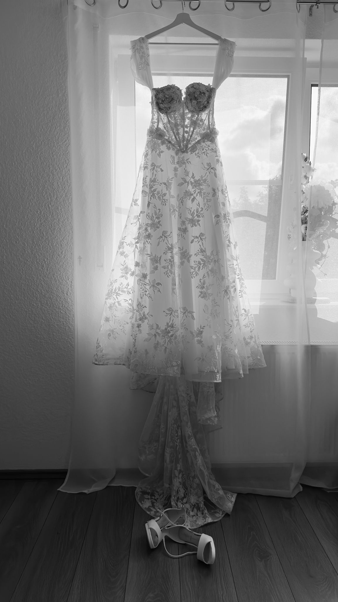 Elegant floral lace wedding dress hanging by window in black and white photo.