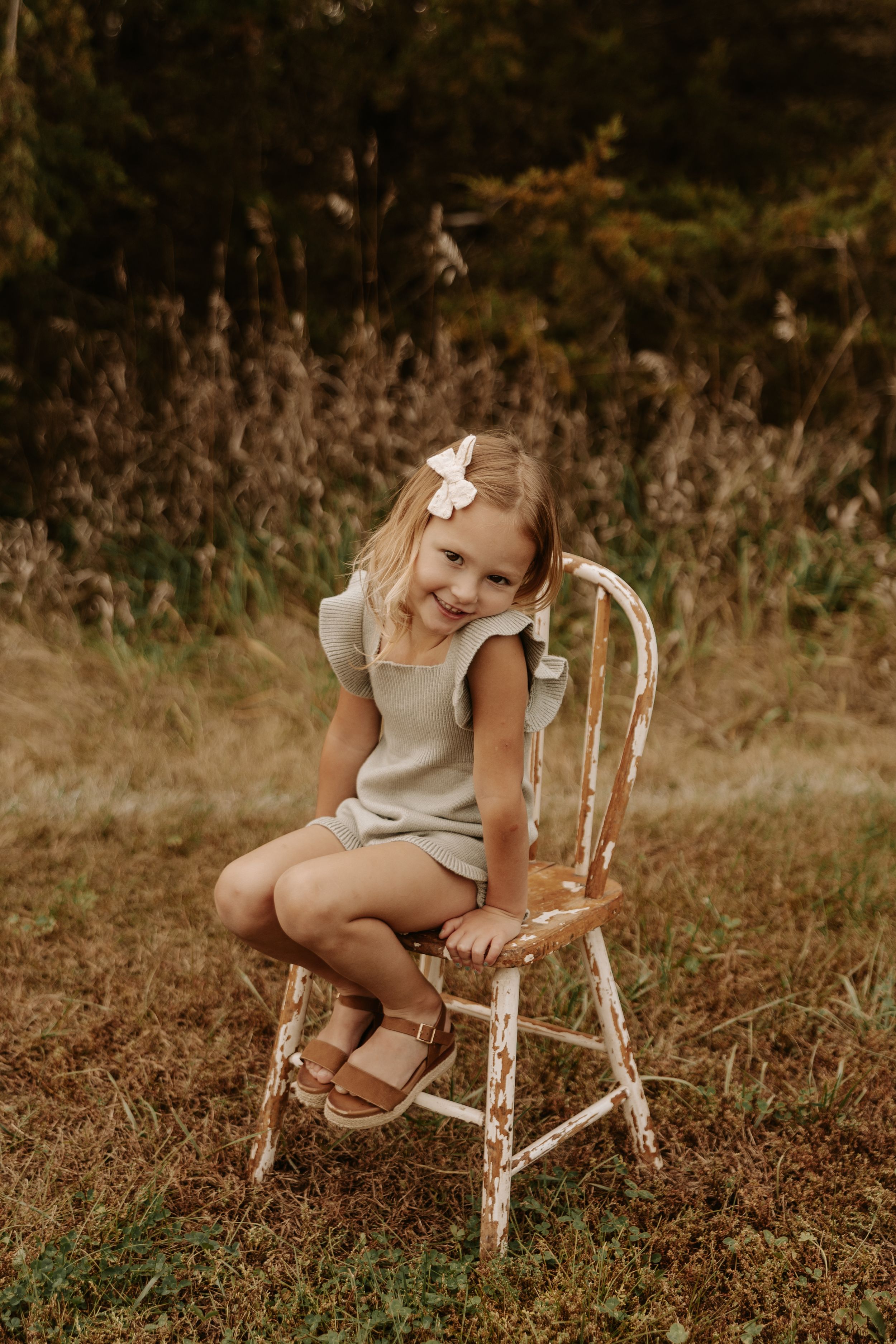 Young girl in sage romper sits on rustic white chair outdoors, smiling with white bow in blonde hair.