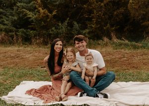Family of four smiling on a white blanket in a grassy field, parents with two young children outdoors.