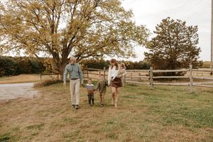 Family of four walking together on a green farm field with wooden fence and autumn trees in background.
