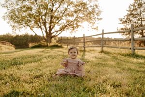 Smiling toddler girl in pink dress sitting on green grass at golden hour near wooden fence.