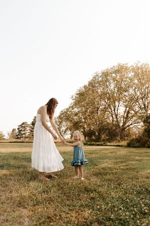 A mother in a white dress holds hands with her toddler daughter in a blue dress in a sunny green field.