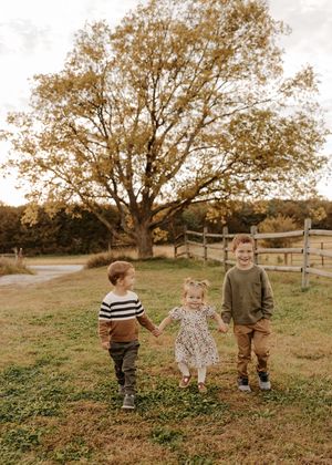 Three young children holding hands in an autumn field with a large golden tree and wooden fence backdrop.