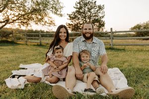 Family of four smiling together on a blanket in a sunny green field at golden hour near a wooden fence.