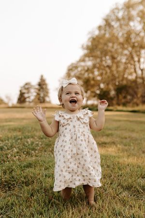 Joyful toddler girl in floral dress with white bow laughing outdoors in green grass field.