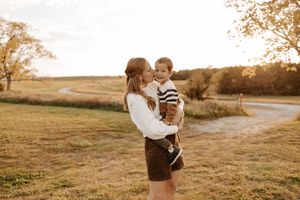 A mother kisses her toddler son while holding him in a sunlit autumn field at golden hour.