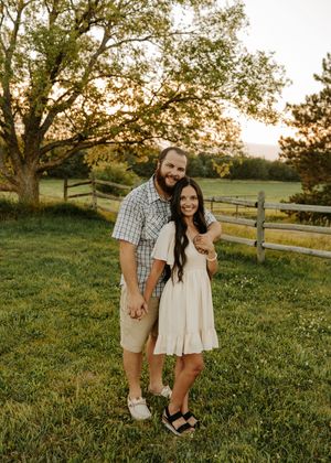 A smiling couple poses together on a lush green farm at golden hour, with a wooden fence and trees in the background.